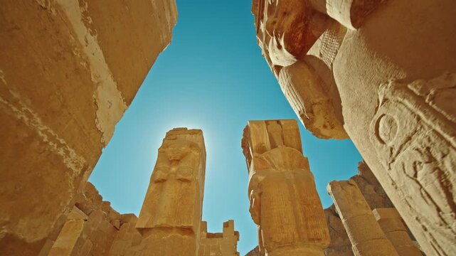 Low angle view of monumental ancient statues with sun flare shining between massive stone pillars at the Temple of Hatshepsut in Luxor, Egypt