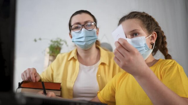 Woman and girl wearing mask study at computer while mother guides homework with book in cozy home setting showing education care and safety during health precaution emphasizing education safety