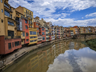 Naklejka premium Colorful houses hanging over the Onyar River in Girona, Spain, reflected in the water under a cloudy blue sky.