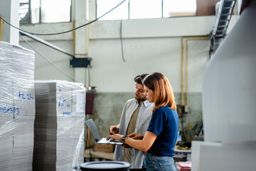 Two professional factory workers use a laptop to manage inventory and production workflow in a printing plant, ensuring high-tech manufacturing quality control and industrial warehouse efficiency