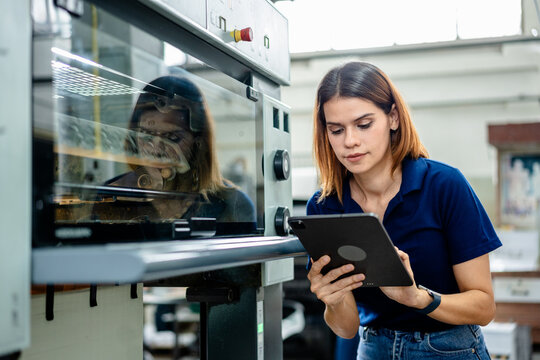 Female worker inspecting machinery in printing factory, using digital tablet for monitoring production and quality control. Concept of manufacturing process, technology, industrial, and efficiency.