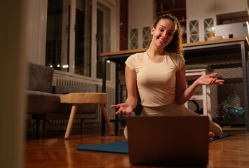 Obraz premium Woman performing a workout at home, sitting on a yoga mat in front of a laptop, smiling and gesturing while participating in an online fitness class in the evening