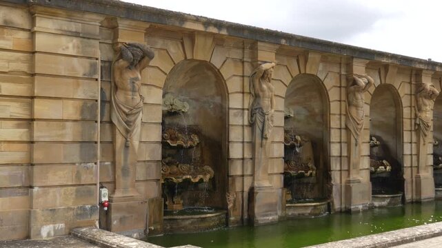 Series of sandstone sculptures supporting arched wall with repeating figures beside stone stairs and green trees.