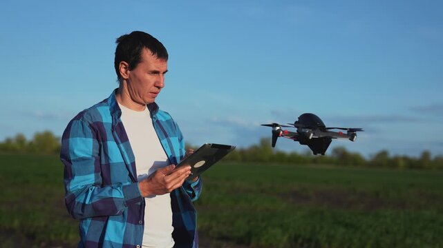 Operating drone with tablet controller pilot man stands in green field controlling black quadcopter drone flight in blue sky using tablet remote for flight test and aerial inspection of land area