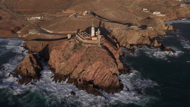 Dramatic aerial view of the Cabo de Gata lighthouse perched on a rugged cliff during a golden sunset