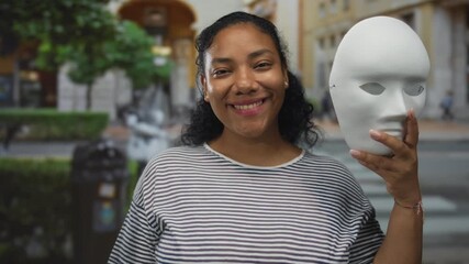 Woman wearing striped shirt smiles broadly and holds white mask with hand on busy city street; joy identity expression.