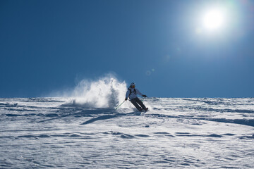 Naklejka premium Skier skiing off piste through fresh powder on a sunny mountain slope, kicking up snow as she descends, creating a dynamic and energetic winter sports action scene under a clear blue sky