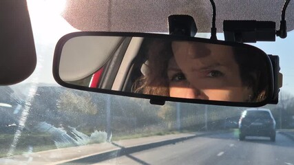 Driving mirror woman, serious female driver reflecting in the rearview mirror while commuting on a suburban road in the morning sunlight.