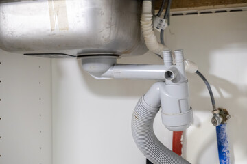 Under kitchen sink showcasing complex gray PVC drain pipes, corrugated hose connection, and water supply lines, essential for plumbing maintenance or installation