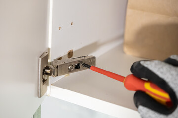 Hand in work glove precisely uses a screwdriver to tighten a metal hinge on a white cabinet, showcasing home improvement and furniture assembly