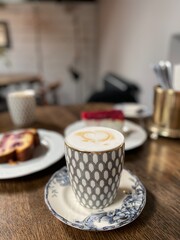 Coffee cup on patterned saucer with latte art on wooden table, background with cake