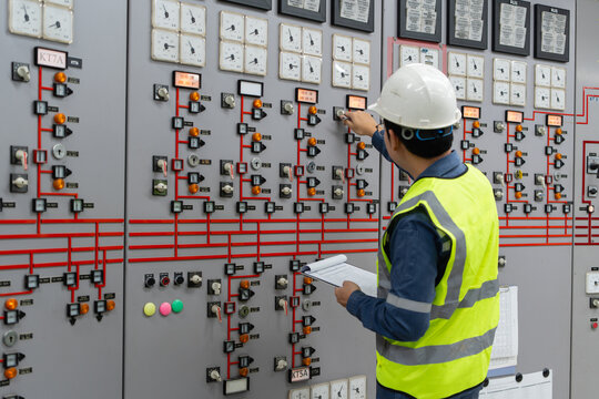 Professional electrical engineer in hardhat and safety vest inspecting control panel and monitoring gauges in power station control room, ensuring industrial energy system safety and efficiency.