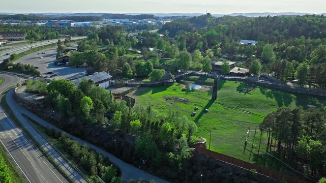 Aerial view of Dyreparken SafariCamp and green fields. Drone footage showing grassy areas, animal enclosures, and nearby buildings in summer with clear skies and lush vegetation.