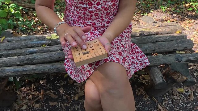 Close-up of woman playing a wooden sliding puzzle while sitting on a bench in the forest. Mindful leisure activity outdoors, slow living, relaxation, focus and calm connection with nature
