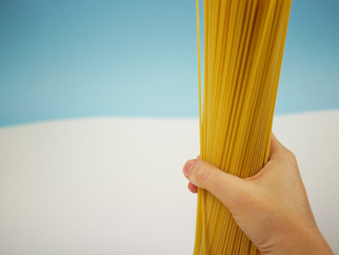 Vertical view of a large bundle of uncooked spaghetti pasta held by a hand on blue and white background, modern food photo of dry Italian ingredients