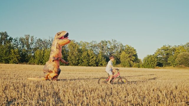 Inflatable T-Rex Chasing Boy Riding Bicycle in Sunny Open Field. Childhood fun.
