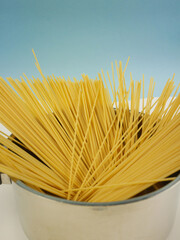 Tall vertical image of uncooked spaghetti rising from a shiny metal saucepan on blue and white background, modern food photo of dry pasta cooking ingredients