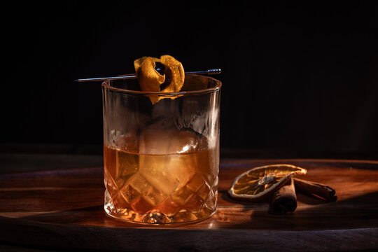 Smoked whiskey cocktail served on the rocks garnished with a dehydrated orange slice and a cherry with dramatic lighting served on a wooden surface.
