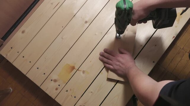 Close-up of a man's hands using a cordless power drill to screw a small wooden board onto a larger wooden surface. Home improvement and DIY carpentry work in progress
