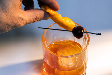 Close up of a hand placing an orange slice in a manhattan cocktail served with one large ice cube on a reflective white surface.