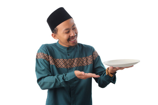 Excited Asian Muslim man wearing traditional Indonesian Muslim clothing, a teal baju koko shirt and black peci cap (songkok), holding and showing an empty white plate, isolated on a white background