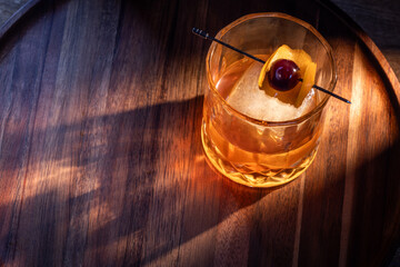 whiskey cocktail served on the rocks garnished with an orange peel anda cherry with dramatic lighting served on a wooden surface.