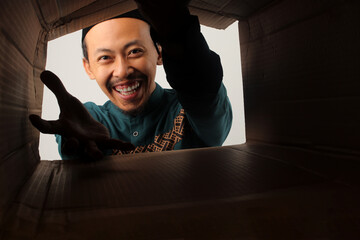 Happy Asian Muslim man unpacking and opening a carton box, looking inside with excitement, symbolizing delivery, surprise, gift, or online shopping, isolated on a white background