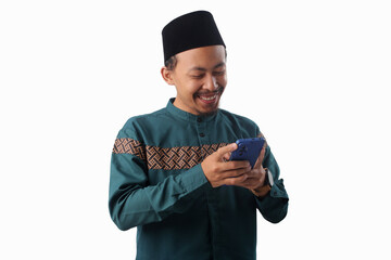 Happy Asian Muslim man in a green traditional outfit smiling while reading good news on his phone. Isolated on white background. Concept of joyful communication, positive updates, or digital happiness