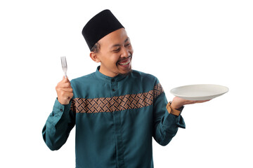 Joyful Indonesian Muslim man wearing a teal koko shirt and black peci (songkok) holding a plain plate and a fork, waiting for iftar to break his fast during Ramadan, isolated on a white background.