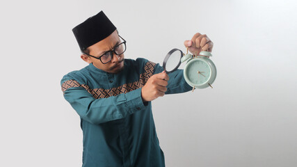 Asian Muslim man in green traditional outfit holding a magnifying glass over alarm clock. Isolated on white background. Time management, activity planning, deadline, and schedule preparation concept
