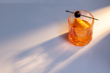 Close up of a manhattan cocktail served with one large ice cube on a reflective white surface with dual lighting tones and high contrast lights adding drama.