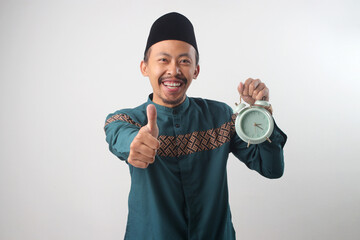 Asian Muslim man in a green traditional outfit giving a thumbs-up gesture to the camera while holding an alarm clock in his hand. Isolated on a white background.