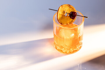 Close up of a manhattan cocktail served with one large ice cube on a reflective white surface with dual lighting tones and high contrast lights adding drama.