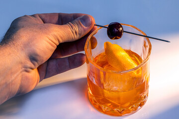 Close up of a hand placing the cherry on top of a manhattan cocktail served with one large ice cube.