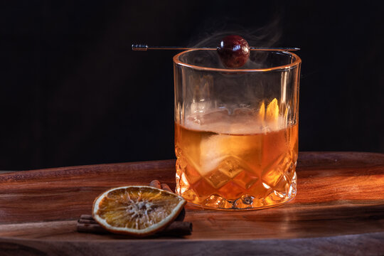 Smoked whiskey cocktail served on the rocks garnished with a dehydrated orange slice and a cherry with dramatic lighting served on a wooden surface.