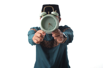 Asian Muslim man in green traditional outfit holding an alarm clock in front of his face. Isolated on white background. Concept of time, identity, or time pressure.