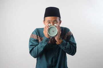 Asian Muslim man in green traditional outfit holding an alarm clock in front of his face, hiding behind it. Suhoor time is almost over during Ramadan fasting. Isolated on white background.