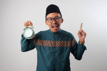 Asian Muslim man in a green traditional outfit pointing at empty copy space while holding an alarm clock in his hand. Isolated on a white background. Concept of time announcement, reminder