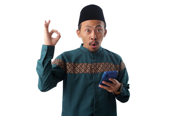 Asian Muslim man in a green traditional outfit showing an OK hand gesture while holding a phone. Isolated on white background. Concept of approval, satisfaction, or successful digital interaction.