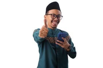 Asian Muslim man in green traditional outfit and eyeglasses giving a thumbs up to the camera while holding a phone. Isolated on white background. Concept of approval, success, or positive experience