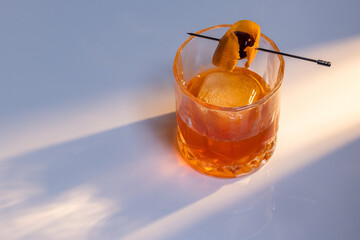 Close up of a manhattan cocktail served with one large ice cube on a reflective white surface with dual lighting tones and high contrast lights adding drama.