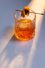 Close up of a manhattan cocktail served with one large ice cube on a reflective white surface with dual lighting tones and high contrast lights adding drama.