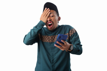 Surprised Asian Muslim man in green traditional outfit covering his face with a humorous expression while reacting to something on his phone. Isolated on white background.