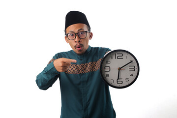 Surprised Asian Muslim man wearing green traditional outfit, pointing at a clock he is holding in his hand. It&rsquo;s time for iftar, the meal to break the fast during Ramadan. Isolated on white background