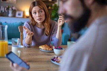 Woman looking disappointed at man on phone during breakfast at home