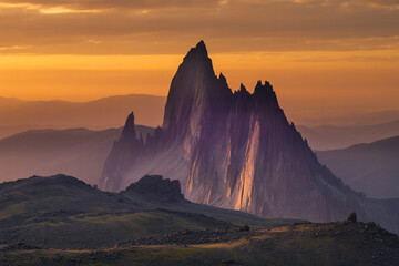 A dramatic landscape photograph of a jagged mountain peak at sunset. The mountain has steep, rocky cliffs and sharp peaks, illuminated by warm golden light from the setting sun.