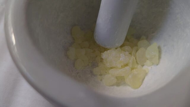 Top view of crushing aromatic Mastic gum resin in a white ceramic mortar, showing the process of grinding translucent yellow resin crystals into a fine powder.
