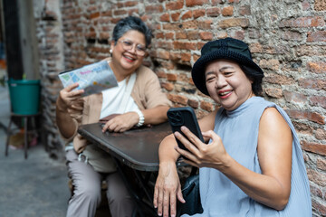 Cheerful senior asian women travelers sitting outside brick wall using smartphone showing photo sharing joyful moment together enjoying retirement lifestyle vacation travel friendship adventure