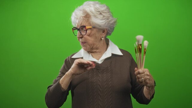Senior woman holding several soft brushes in one hand while pinching her nose with the other in a green screen studio, looking away; disgust.