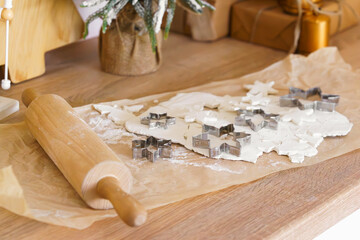 Festive Christmas baking process with rolled dough, metal cookie cutters for gingerbread and wooden rolling pin on parchment paper. Holiday kitchen scene preparing homemade cookies for celebrations.